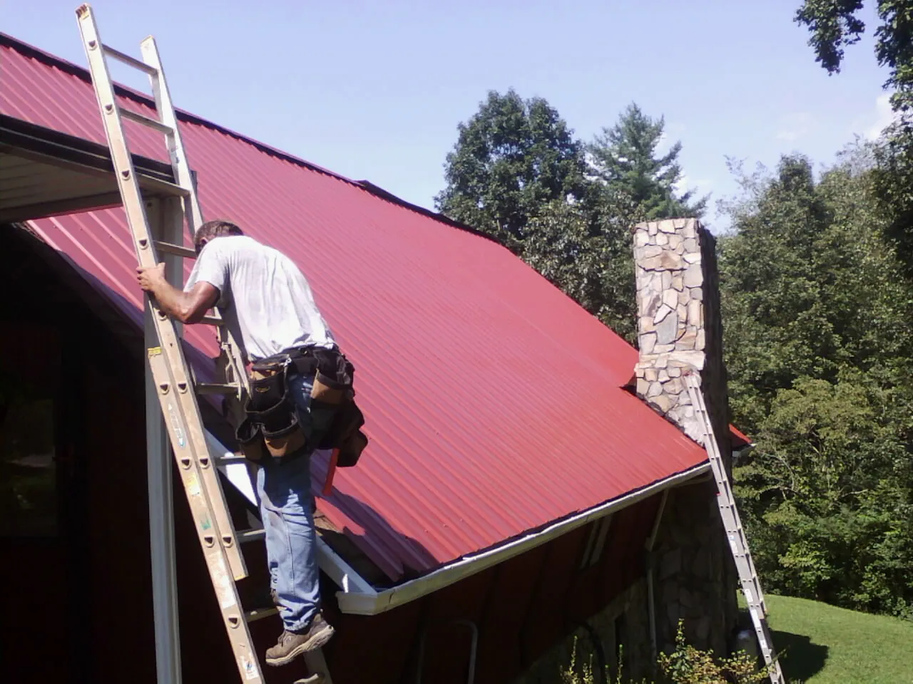 Roofer on a ladder beside a red metal roof and stone chimney — Soffit & Fascia Repair by Roofing Brothers in Bonney Lake
